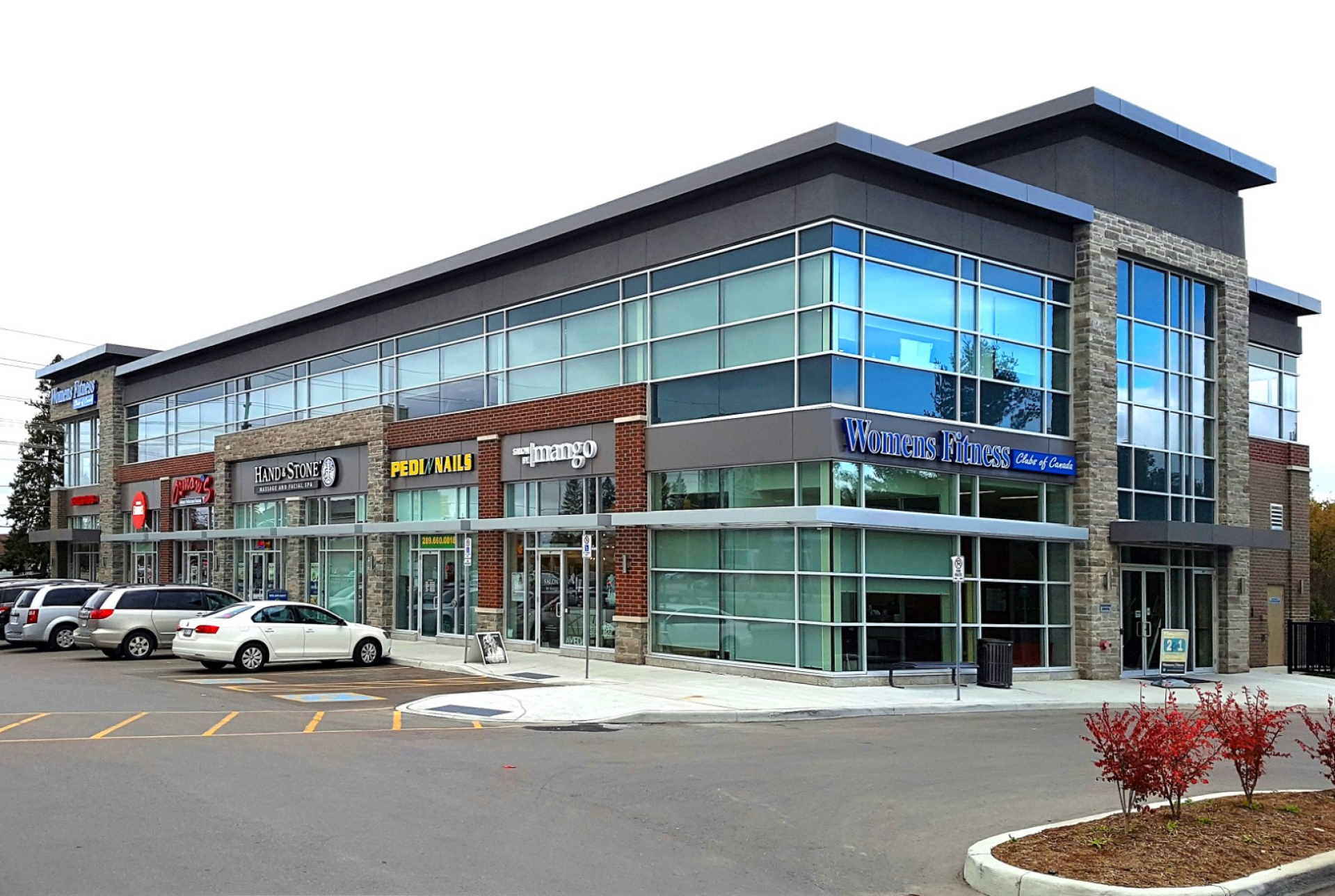 A modern two‑storey retail plaza with large glass storefronts, mixed brick and metal exterior finishes, and visible shop signage. Cars are parked along the curb in front of the building under an overcast sky.